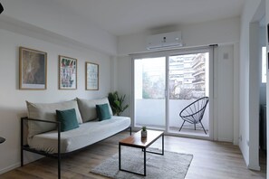 Una sala de estar moderna con un sofá, una mesa de centro y una silla. Hay cuadros enmarcados en la pared y un ventanal con vistas a edificios.
