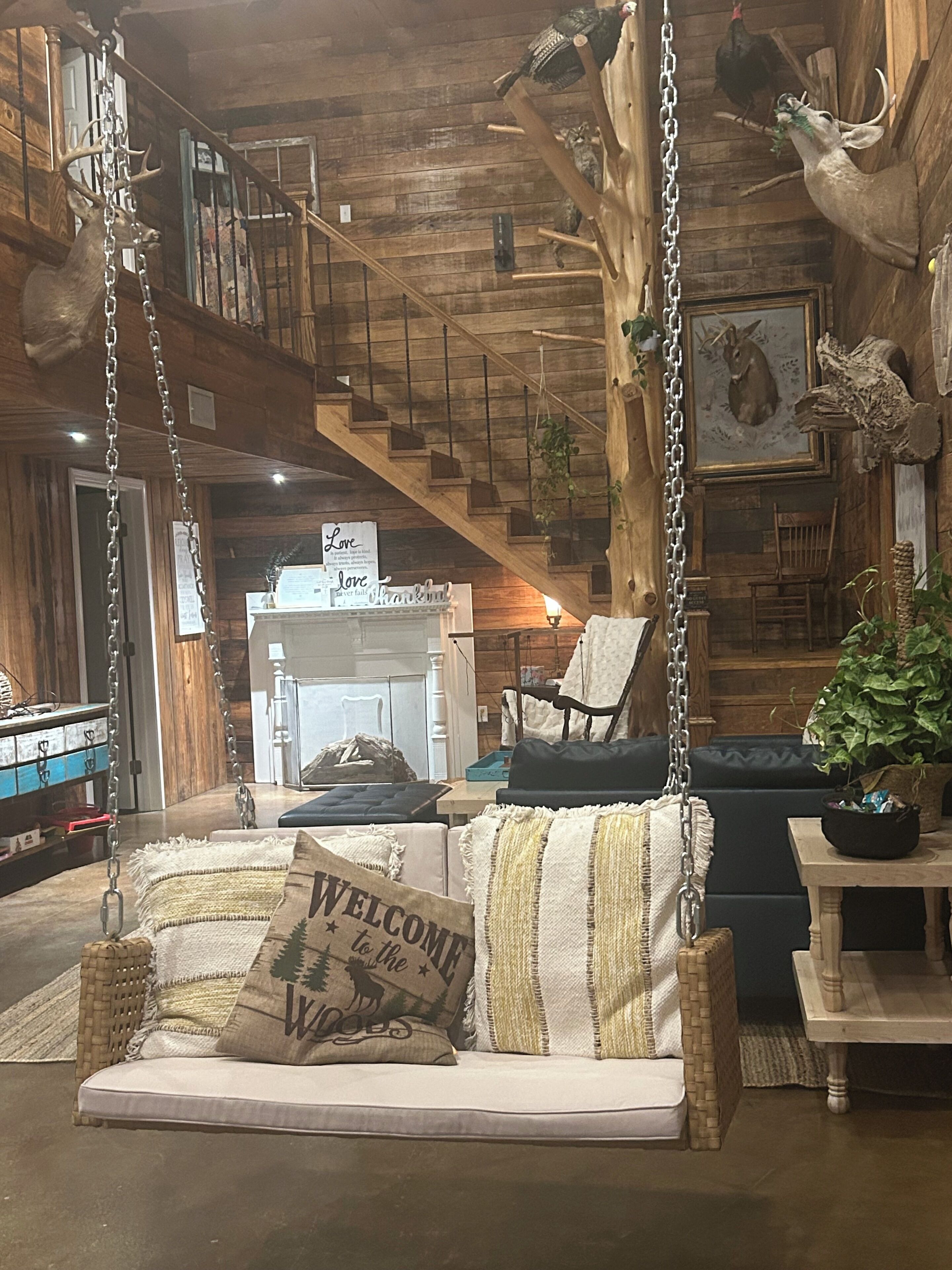 A rustic interior with a wooden staircase, a hanging swing chair, a sofa with a 'Welcome to the Woods' pillow, and a rustic wall decor.. Serene 2-bedroom bed & breakfast four miles from Lake Okhissa in Meadville
