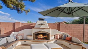 An outdoor seating area with a stone fireplace, cushioned benches, and an umbrella. - Play in Paradise with 20 Pals: Pool, Mini Golf, Pickleball,  Spanish Flair (Scottsdale)