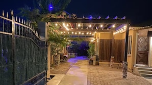 Un chemin pavé mène à une entrée abritée, illuminée de guirlandes lumineuses, ainsi qu’à un espace délimité par une clôture décorative et agrémenté de plantes en pot.