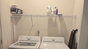 A laundry room with two front-loading washers and dryers, a metal shelf above holding cleaning supplies, and a fire extinguisher on the left. - Relaxing Small Town Getaway in Bright 3B/3.5 Ba TH in Wendell (Wendell)
