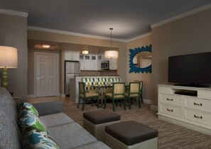 Un salon comprenant un canapé, des poufs, un téléviseur, un coin repas avec des chaises et une table, et une cuisine en arrière-plan.