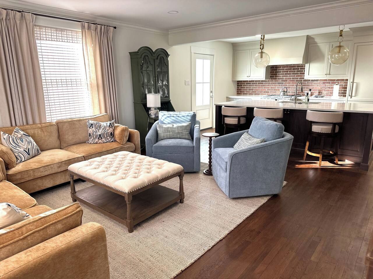 A living room with a sofa, ottoman, and chairs, a kitchen with a brick backsplash, and a dining area with bar stools.