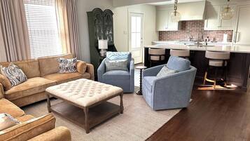 A living room with a sofa, ottoman, and chairs, a kitchen with a brick backsplash, and a dining area with bar stools.