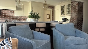 Uma sala de estar moderna com duas poltronas azuis, um sofá bege e uma cozinha ao fundo.