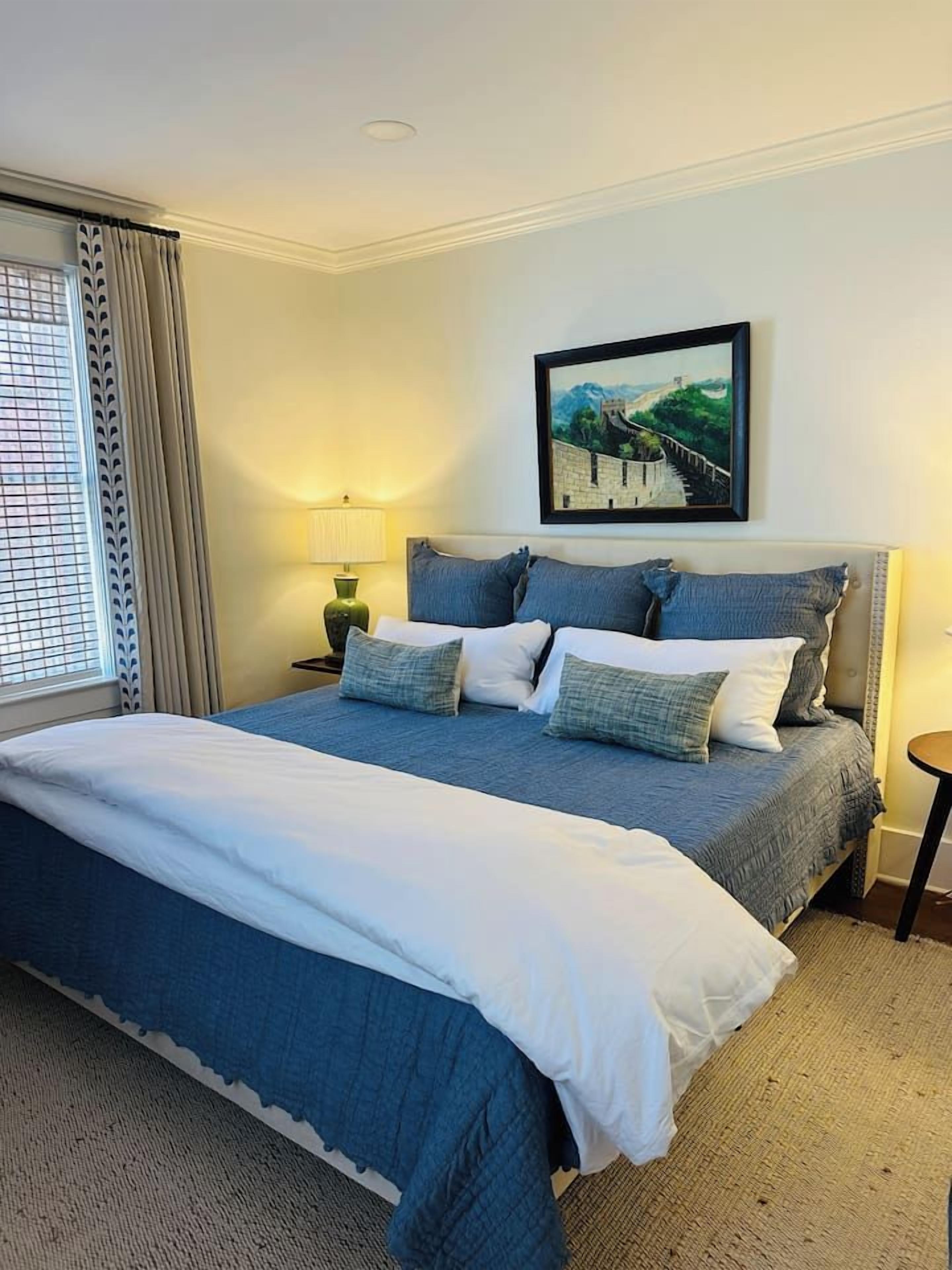 A neatly made bed with a blue and white duvet, a framed picture of a bridge on the wall, and a lamp on a bedside table.