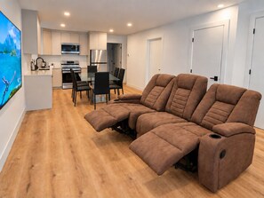 En moderne stue med en hjørnesofa, et spiseområde med bord og stole, samt et køkken med hårde hvidevarer.