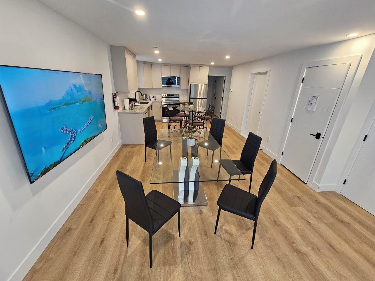 Una cocina moderna con armarios blancos, un refrigerador de acero inoxidable y una zona de comedor con una mesa de vidrio y sillas negras.