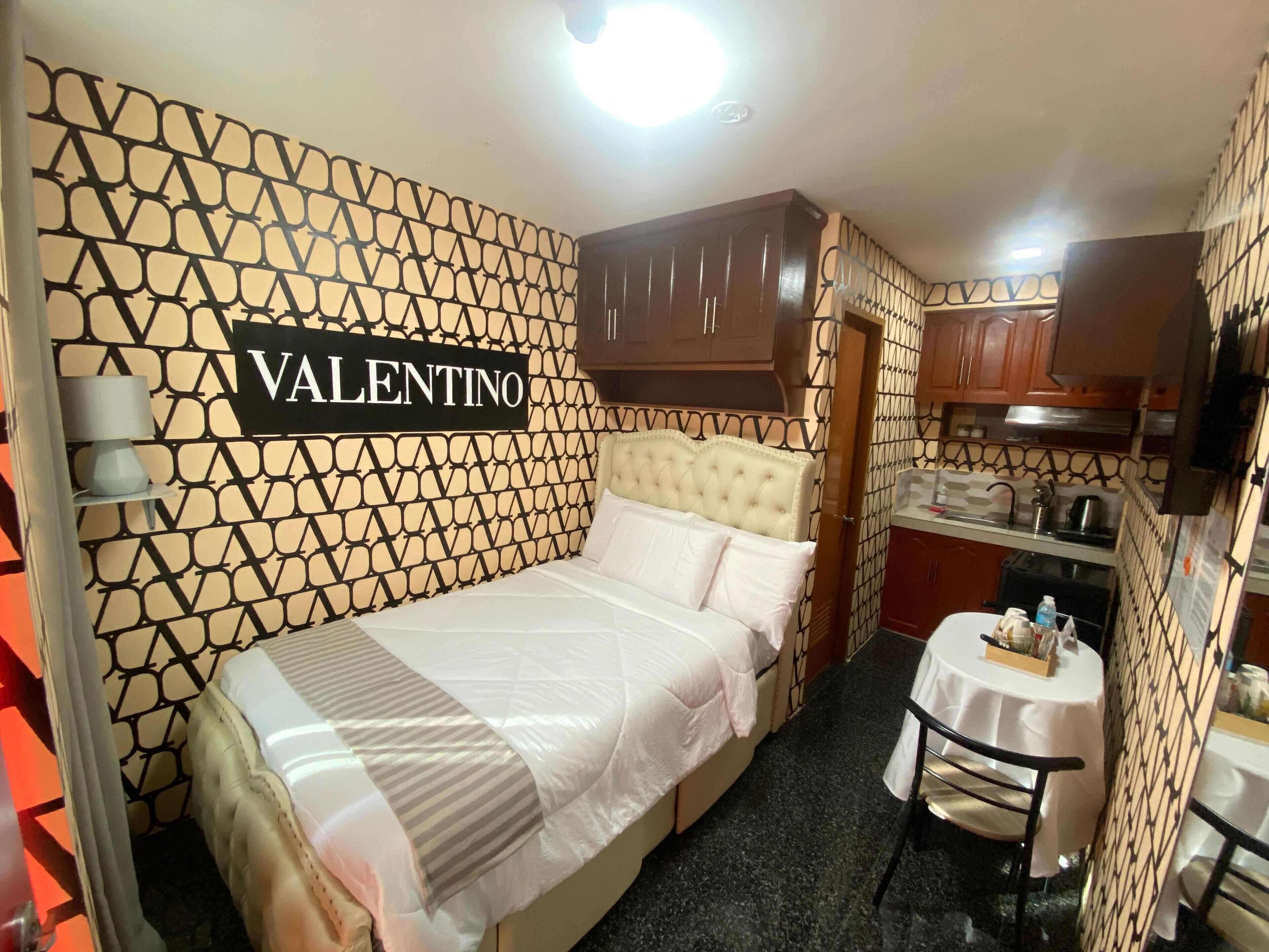 Une chambre à coucher avec un lit, une coiffeuse avec un miroir et une enseigne sur laquelle on peut lire « VALENTINO ».