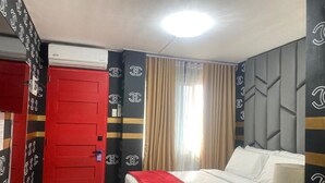 A hotel room with a red door, a bed with white bedding, a red throw blanket, a bedside table with a lamp, and a window with curtains.