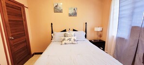 A bedroom with a bed, a nightstand, a lamp, and two wall-mounted paintings. - 2-BR apt in beautiful St Thomas filled with local flava (Charlotte Amalie)