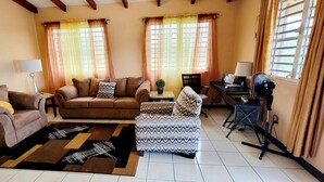 Een woonkamer met een bank, fauteuil, voetenbankje, een bijzettafeltje met een lamp en een ventilator.