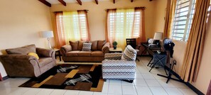 Un salon comprenant un canapé, un fauteuil et un pouf, une petite table avec une lampe et un ventilateur.