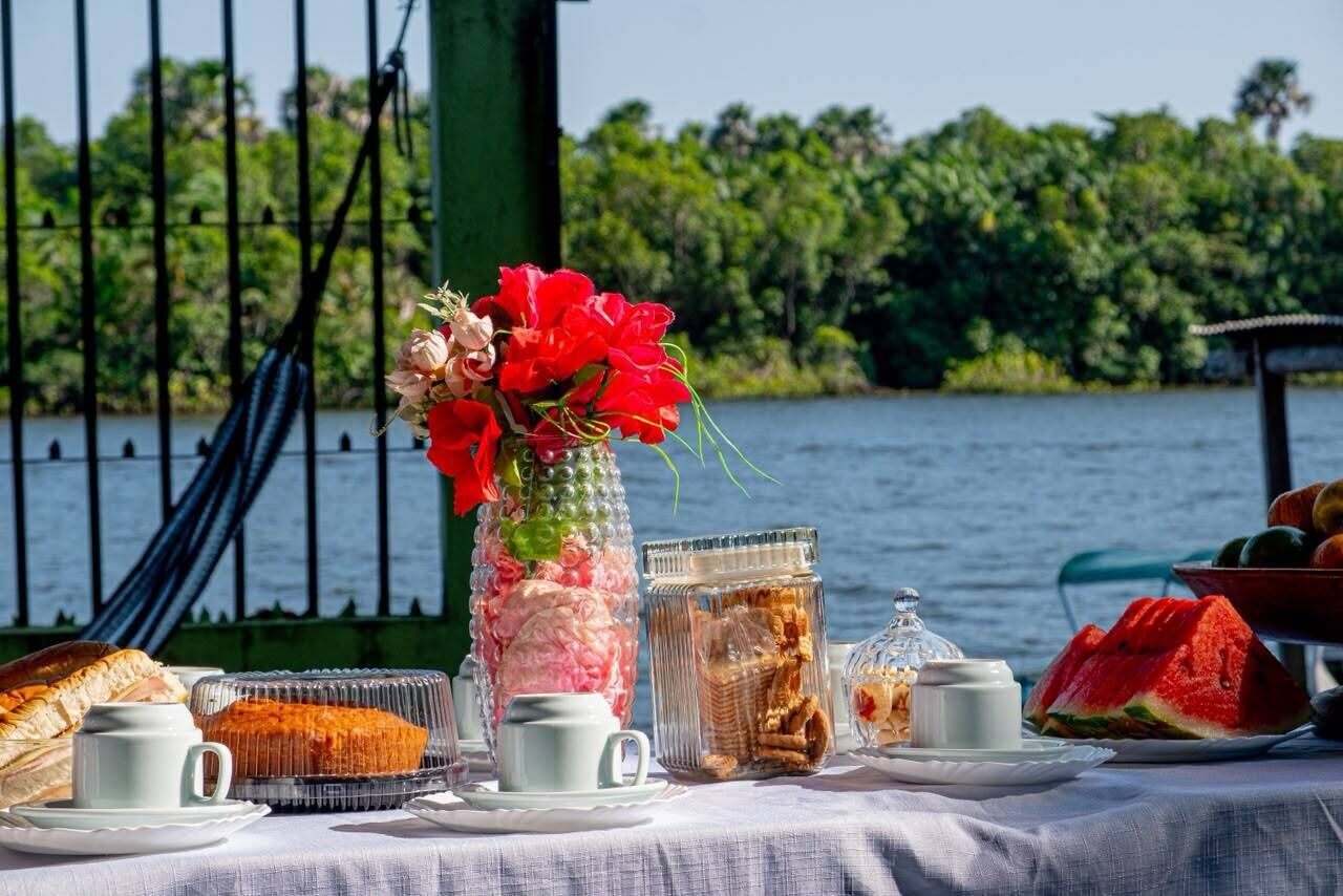 A table set with food and drinks, a vase with flowers, and a view of a body of water and greenery.