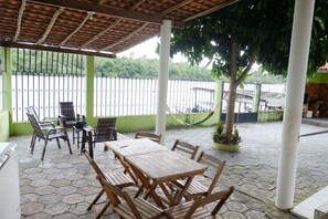 Eine überdachte Terrasse mit sechseckigem Steinboden, Holztischen und -stühlen, einer Hängematte und Blick auf ein Gewässer.