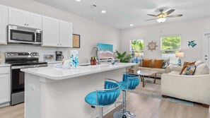 A modern kitchen with a central island, a dining area with bar stools, a living room with a sofa, and a ceiling fan. - Light on Lumen Beach Retreat (Panama City Beach)