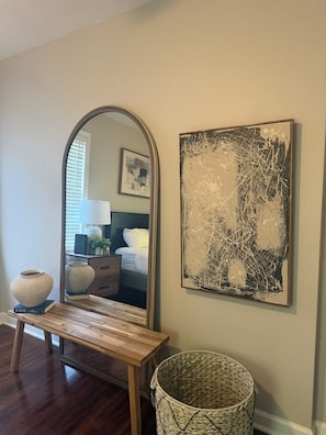A room with a wooden bench, a large mirror, a wall-mounted abstract painting, a wicker basket, and a bed with a nightstand.