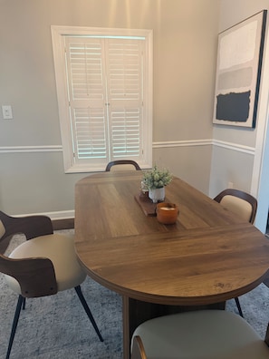 A dining room with a large wooden table, chairs, a potted plant, and a window with blinds. - Clark Retreats (Atlanta)