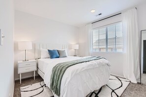 Een slaapkamer met een bed, nachtkastjes, lampen, een raam met jaloezieën en een deur.