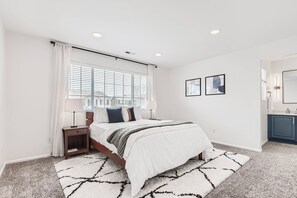 Ein Schlafzimmer mit Bett, Nachttischen, einem Fenster mit Jalousien, einem Teppich und einem Badezimmer mit Spiegel und Waschbecken.