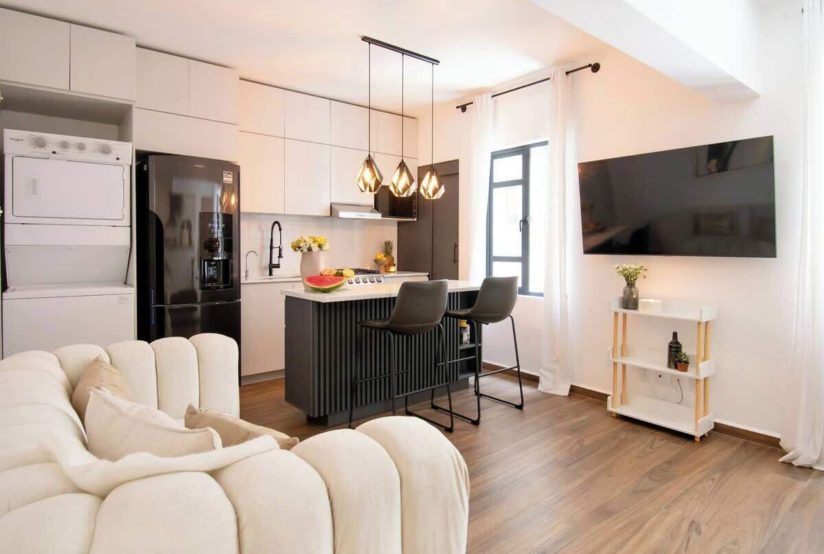 Une cuisine moderne dotée d’un îlot central, d’un coin repas, d’un espace de vie avec un canapé et d’un téléviseur fixé au mur.