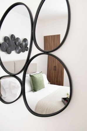 Ein Schlafzimmer mit einem Bett, Kissen, einem grünen Kissen, einem hölzernen Kleiderschrank und einem an der Wand befestigten Spiegel, der den Raum reflektiert.