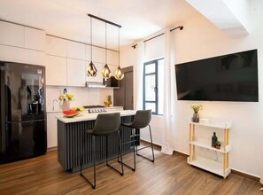 Eine moderne Küche mit einem schwarzen Kühlschrank, einer zentralen Kochinsel mit Barhocker, einem weißen Sideboard und einem wandmontierten Flachbildfernseher.