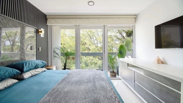 A bedroom with a bed, a bedside table, a TV, and a large window with a view of trees. - Massive rooftop terrace steps from everything (Mexico City)
