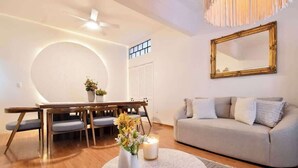 Ein Wohnzimmer mit rundem Couchtisch, einer Couch, einem Esstisch mit Stühlen, einem Deckenventilator und einem großen Spiegel an der Wand.