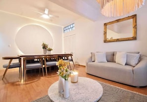 Un salon comprenant une table basse ronde, un canapé, une table à manger avec des chaises, un ventilateur de plafond et un grand miroir au mur.