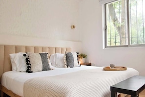 Une chambre avec un grand lit, une table de chevet, une fenêtre donnant sur des arbres, et un plateau avec une tasse et une pâtisserie posés sur le lit.