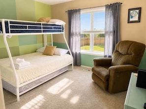 A bunk bed with pillows, a brown armchair, a window with curtains, and a framed picture on the wall.
