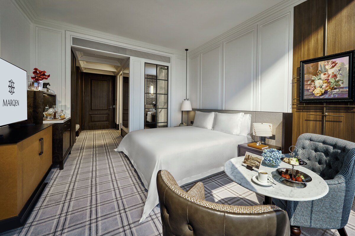 A hotel room with a bed, a desk with a lamp, a chair, a small table with a tray of snacks, and a painting on the wall.