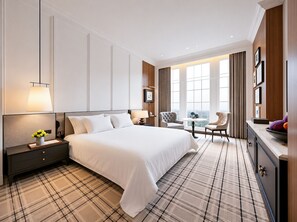 A hotel room with a large bed, a nightstand with a lamp, a chair, a small table, and a view of the city through large windows. - Marqen Hotel (West Jakarta)