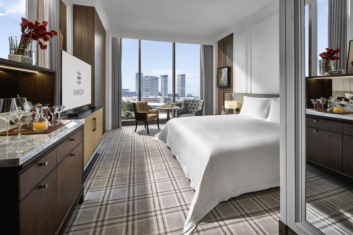 Habitación de hotel con cama, escritorio con minibar, vista panorámica de la ciudad y un ventanal grande.