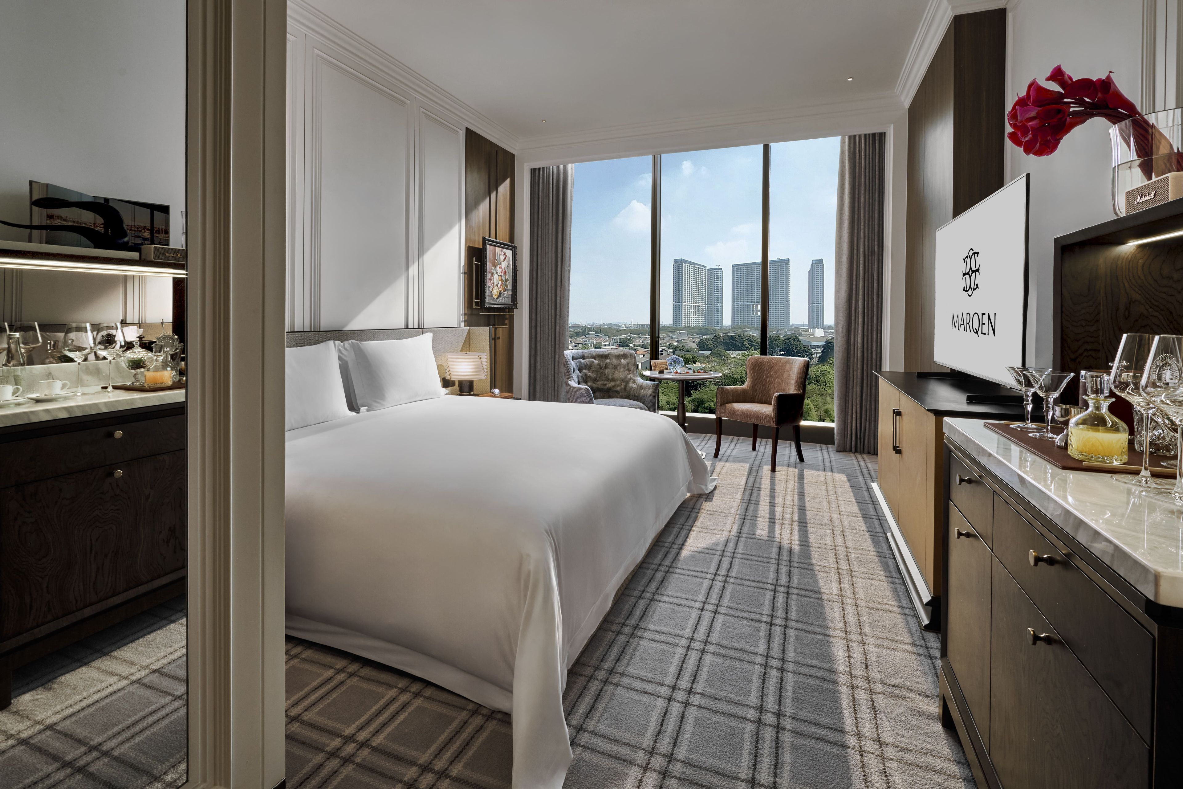 A hotel room with a large bed, a desk with a TV, a view of the city, and a dining area with a table and chairs.