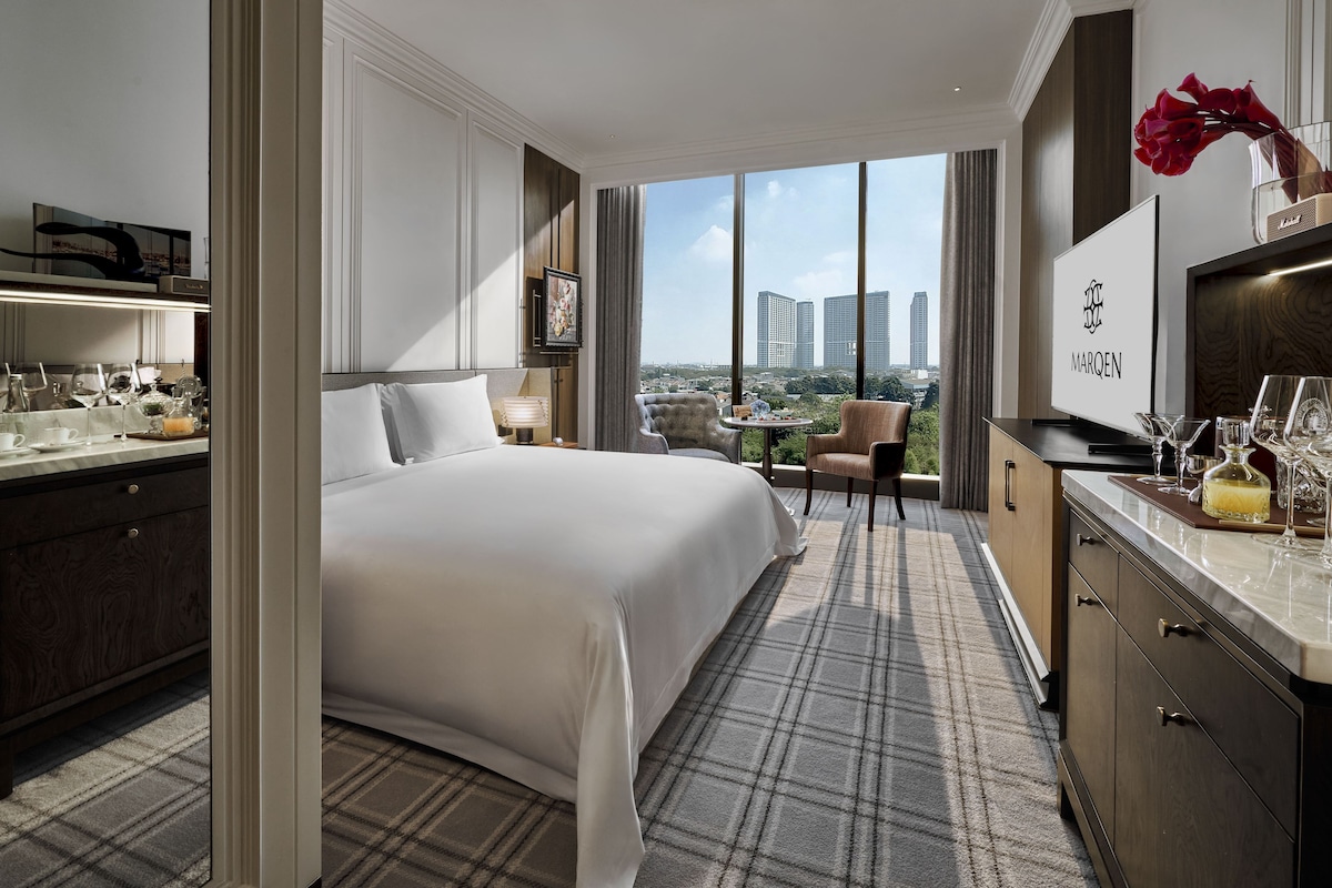 Habitación de hotel con una cama grande, un escritorio con televisor, vistas de la ciudad y una zona de comedor con mesa y sillas.