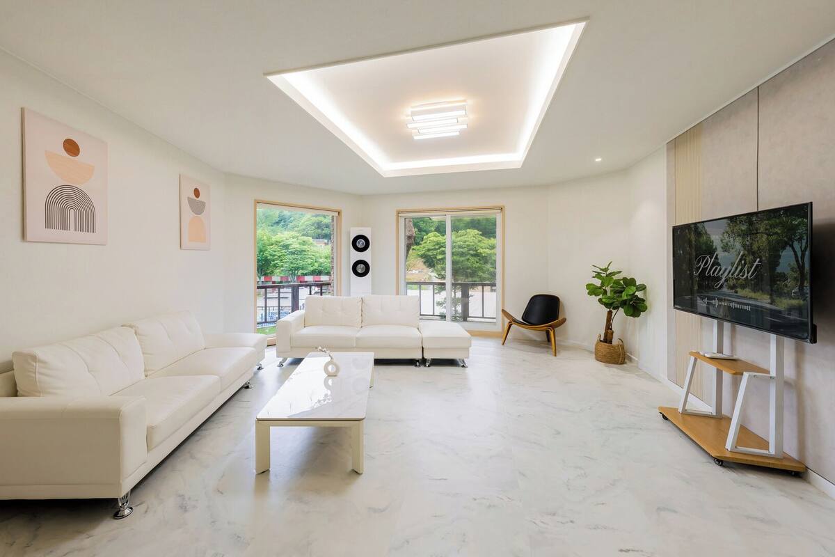 Una sala de estar moderna con un sofá blanco, una mesa de centro, un televisor sobre un mueble bajo y un ventanal grande.