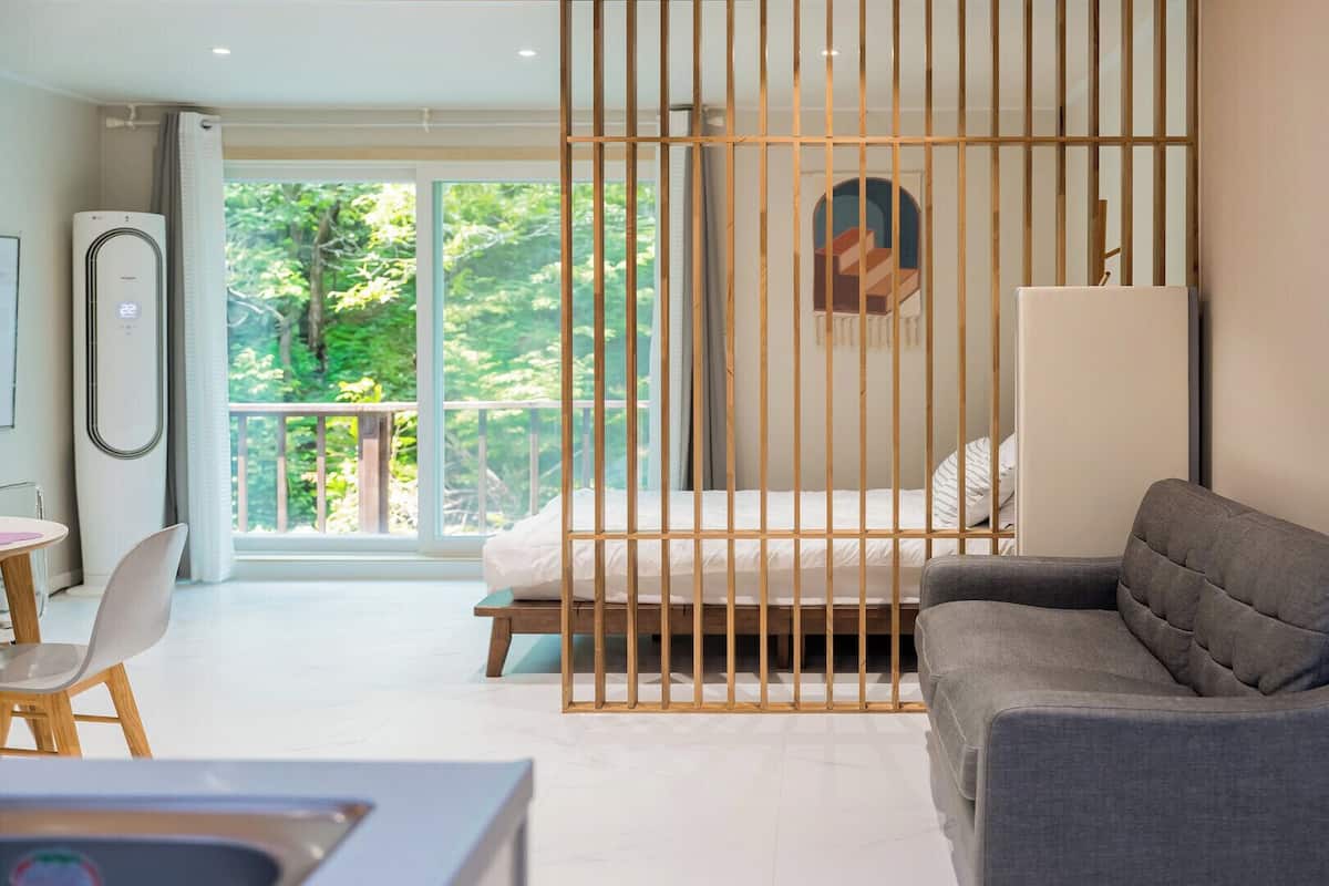 Habitación moderna con cama, sofá gris, escritorio y ventana grande con vistas a zonas verdes.