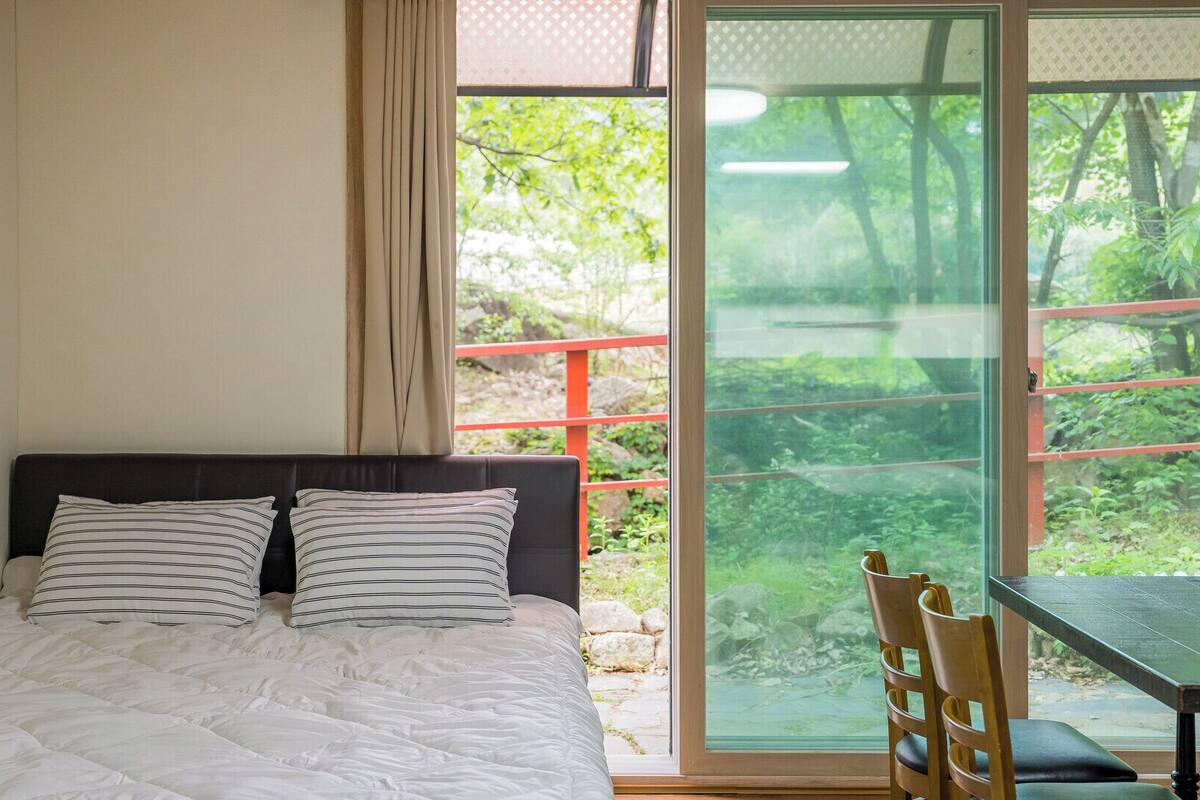 Una cama con edredón blanco y almohadas a rayas, una ventana con vistas a la vegetación y una mesa con una silla afuera.
