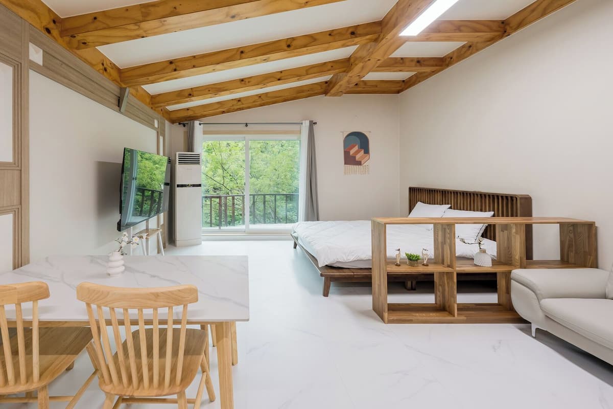 Una habitación con techo de madera, una cama, un televisor, una mesa con sillas y vistas a zonas verdes.