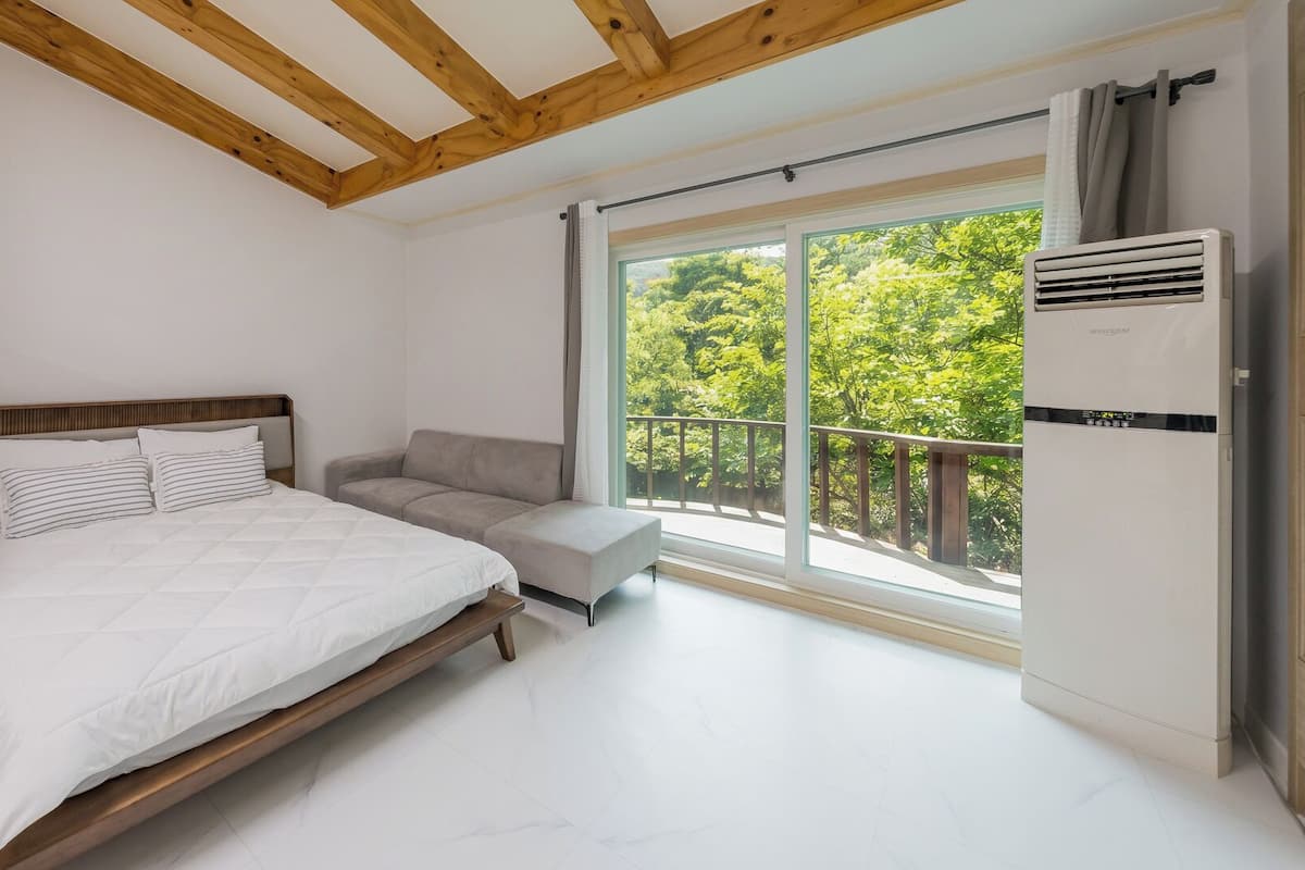Un dormitorio con cama, sofá, ventana con vistas a árboles y un aire acondicionado blanco.