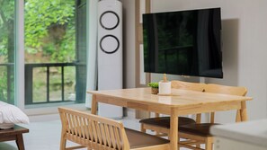 A modern room with a wooden dining table, two chairs, a TV on the wall, and a window with a view of greenery.