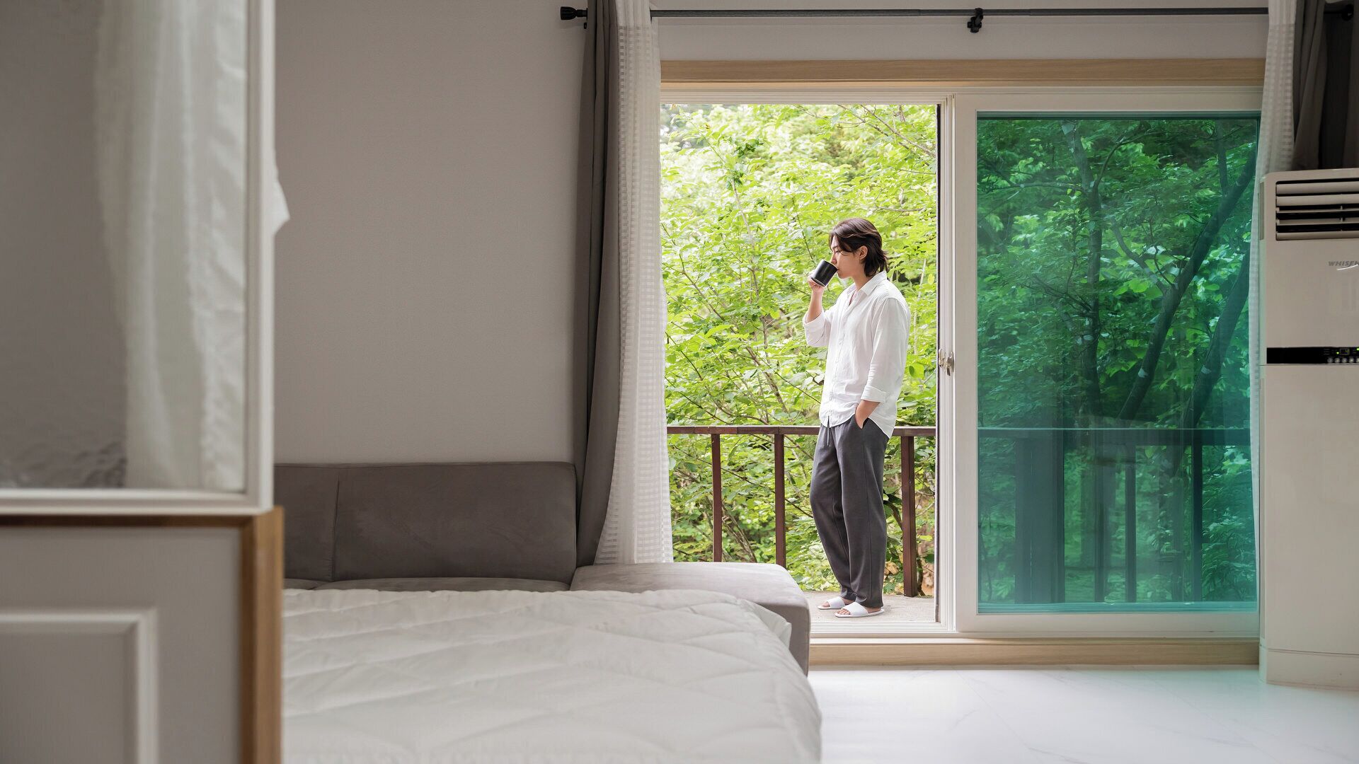 Une chambre avec un grand lit, un balcon donnant sur la verdure et un climatiseur de fenêtre.