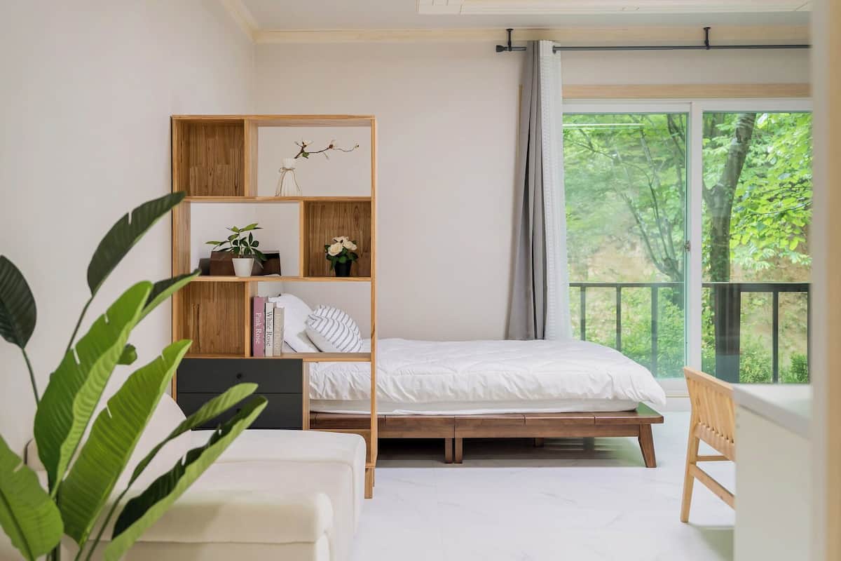 Un dormitorio con cama, estantería de madera, ventana con vista a follaje y una planta en maceta.