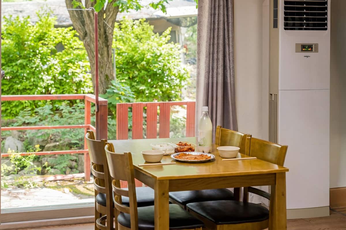 Un comedor con una mesa lista para dos personas, una ventana con vista a follaje y un equipo de aire acondicionado fijado a la pared.