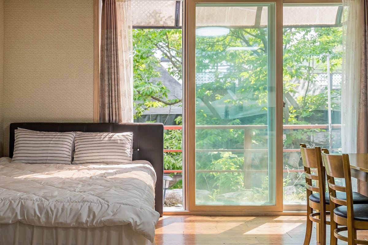 Un dormitorio con cama, una ventana que da a zonas verdes y una zona de comedor con sillas.