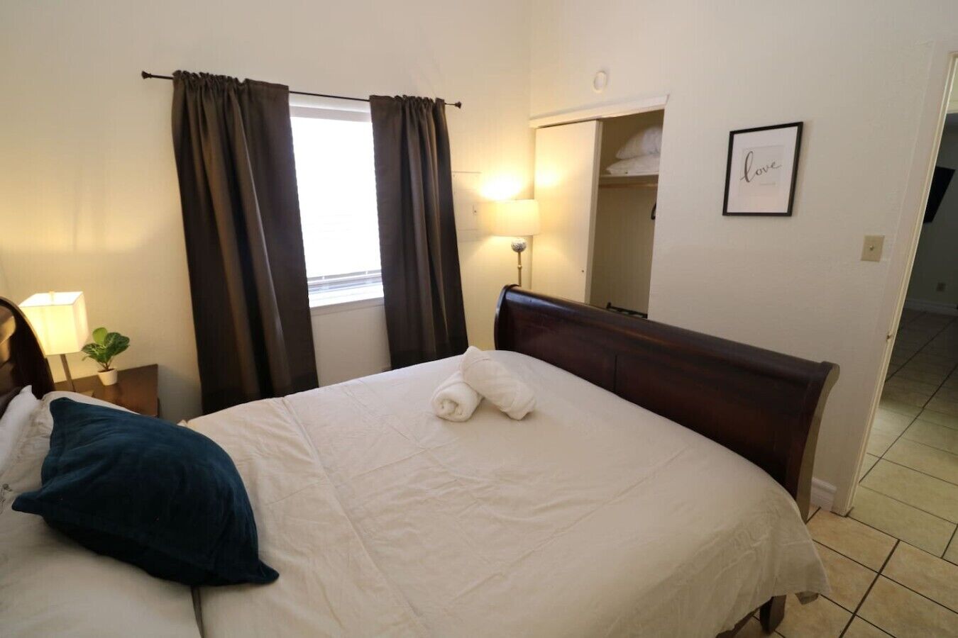 Una habitación de hotel con una cama, una mesita de noche con lámpara, una ventana con cortinas, un espejo y un cuadro en la pared.