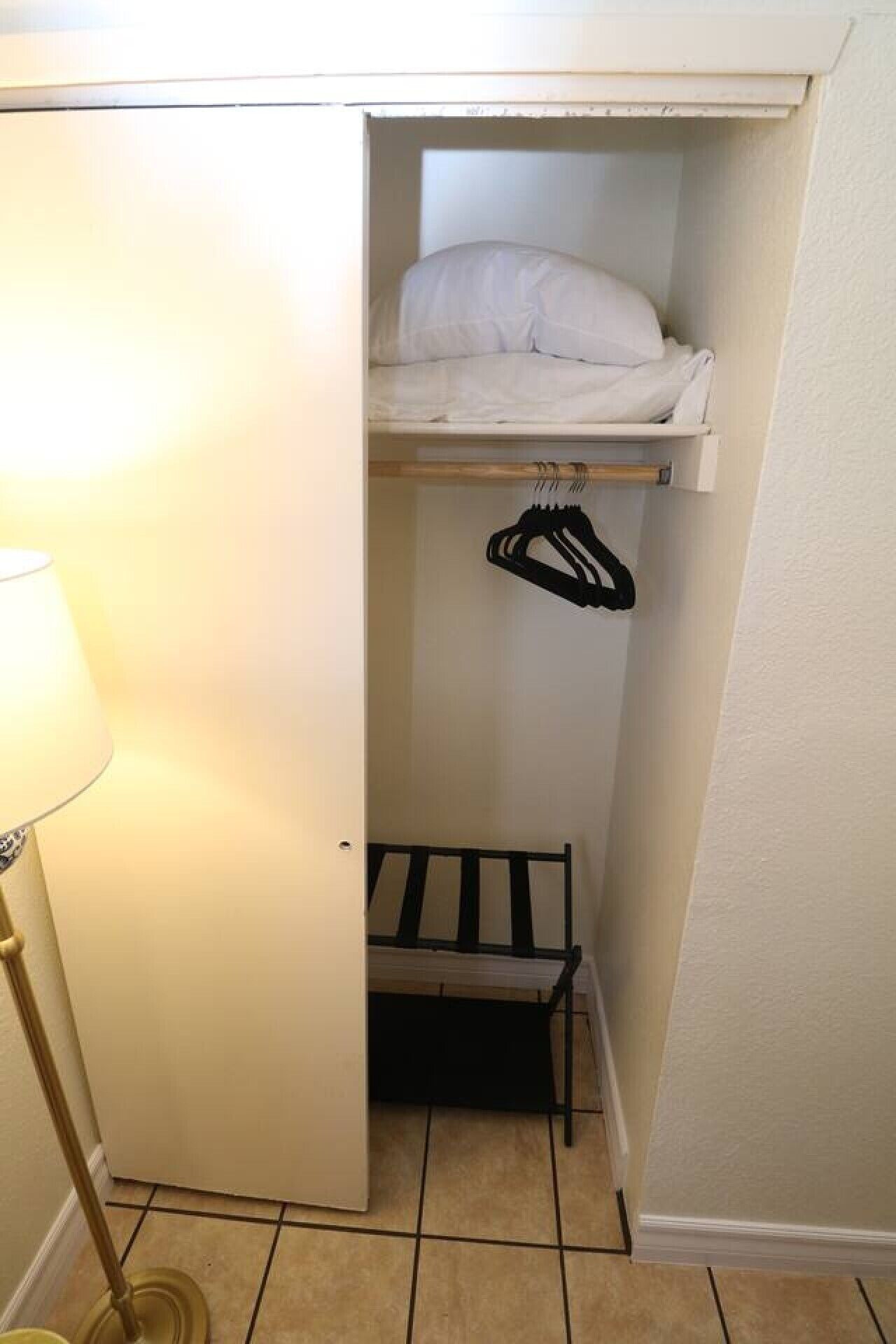 Un petit placard avec une tringle pour suspendre les vêtements, une étagère avec des vêtements pliés et une lampe.