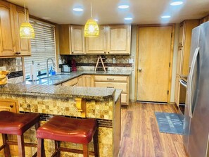 A kitchen with wooden cabinets, a granite countertop, a stainless steel refrigerator, and a bar stool.